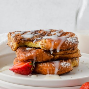 stack of three slices of churro french toast with a bite out of the top slice garnished with white glaze, powdered sugar and strawberry slices with glass of milk in background.