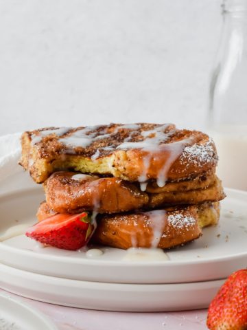 stack of three slices of churro french toast with a bite out of the top slice garnished with white glaze, powdered sugar and strawberry slices with glass of milk in background.