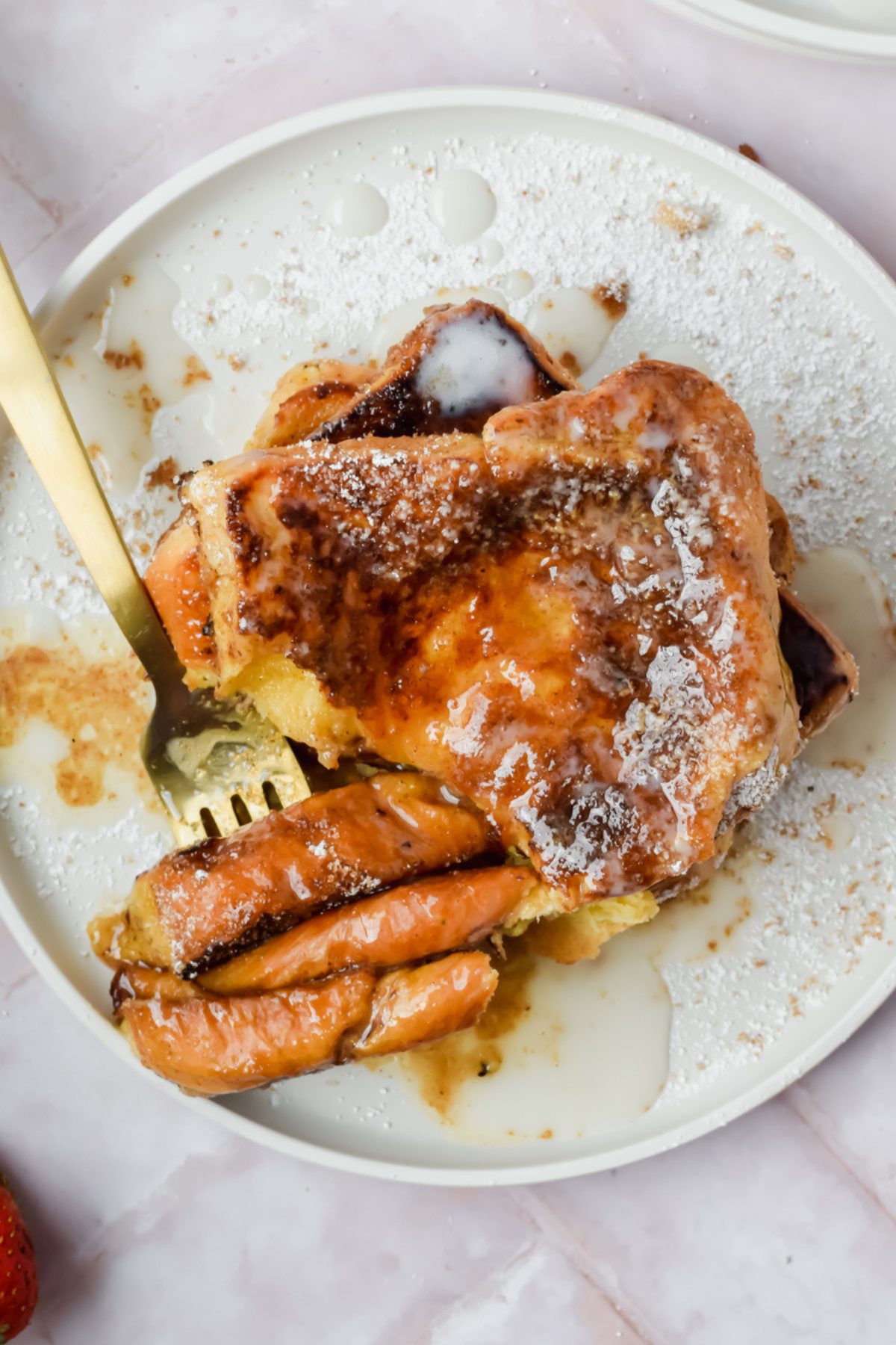 stack of three slices of churro french toast with pieces pierced by a gold fork garnished with white glaze, powdered sugar and strawberry slices with glass of milk in background.