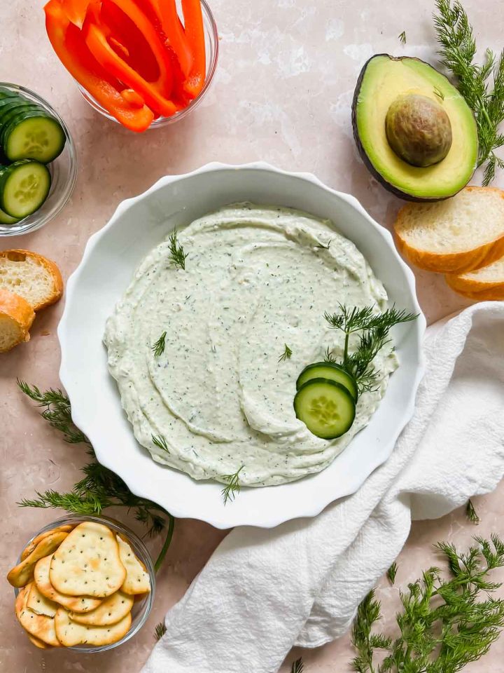 dill feta dip in a white ceramic bowl garnished with fresh dill and cucumbers surrounded by other ingredients on stone background.