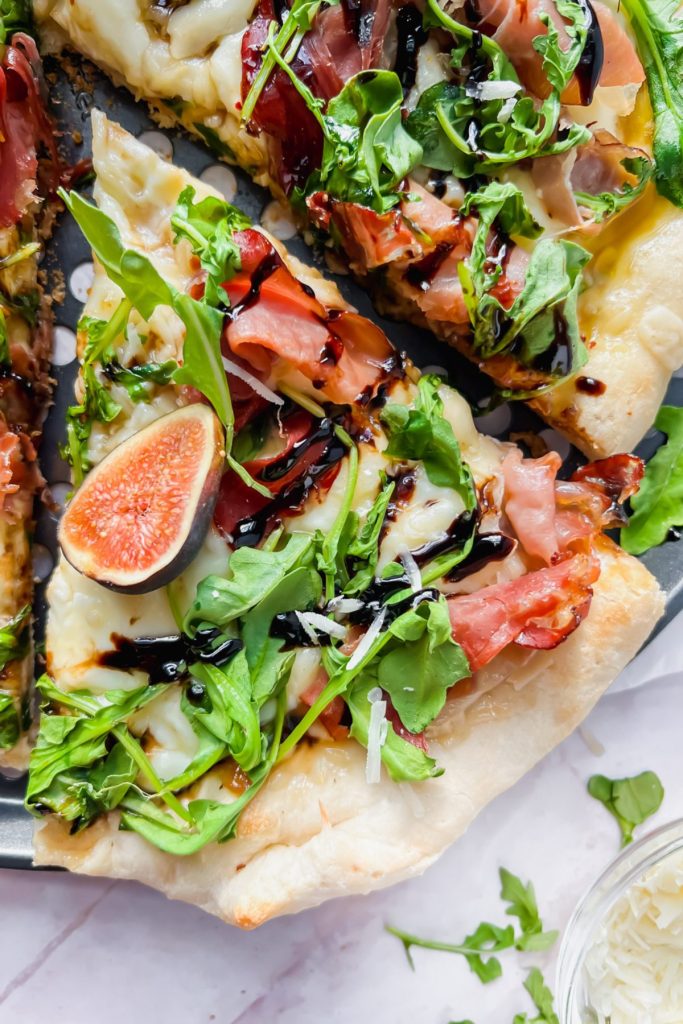 slice taken out of whole fig prosciutto pizza topped with arugula and balsamic drizzle.