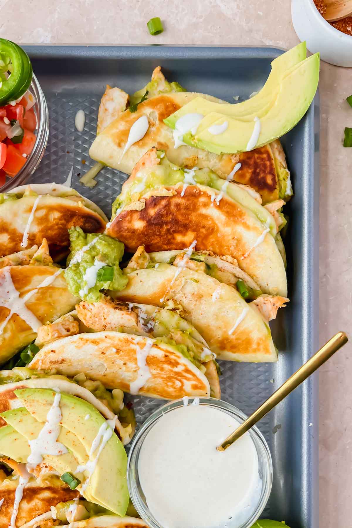 mini quesadillas lined up on gray sheet pan drizzled with guacamole, crema, and garnished with green onions.