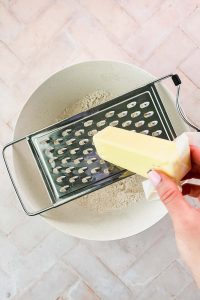 butter stick being grated into bowl of oat flour biscuits dry ingredients.