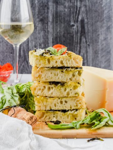 stack of 5 slices of focaccia on wood cutting board with fresh basil surrounding, beside block of parmesan and glass of white wine.