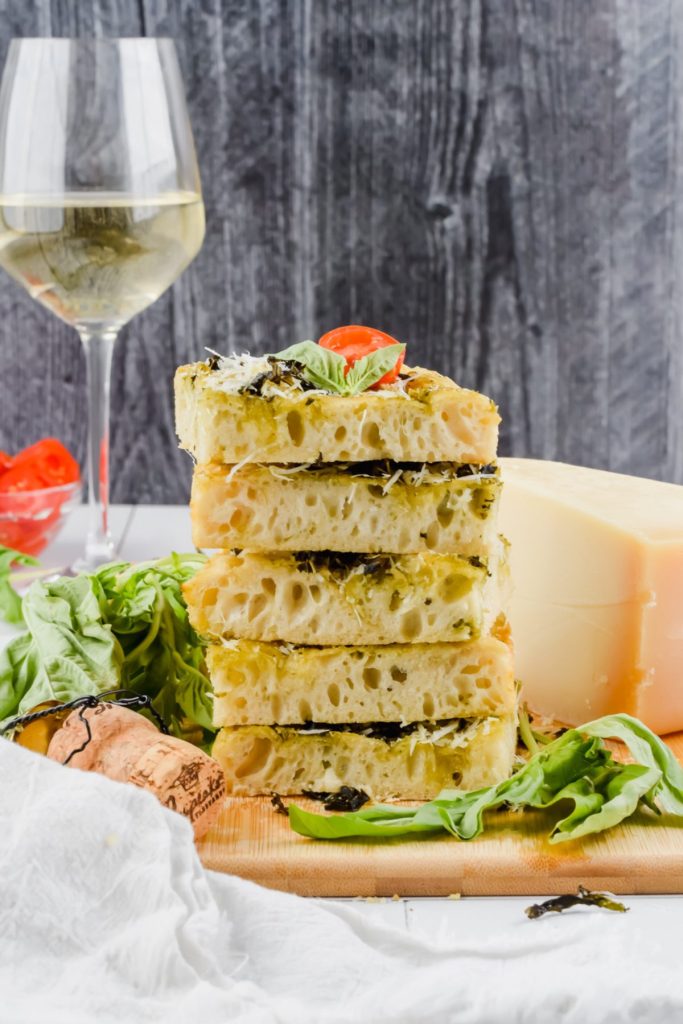 stack of 5 slices of focaccia on wood cutting board with fresh basil surrounding, beside block of parmesan and glass of white wine.