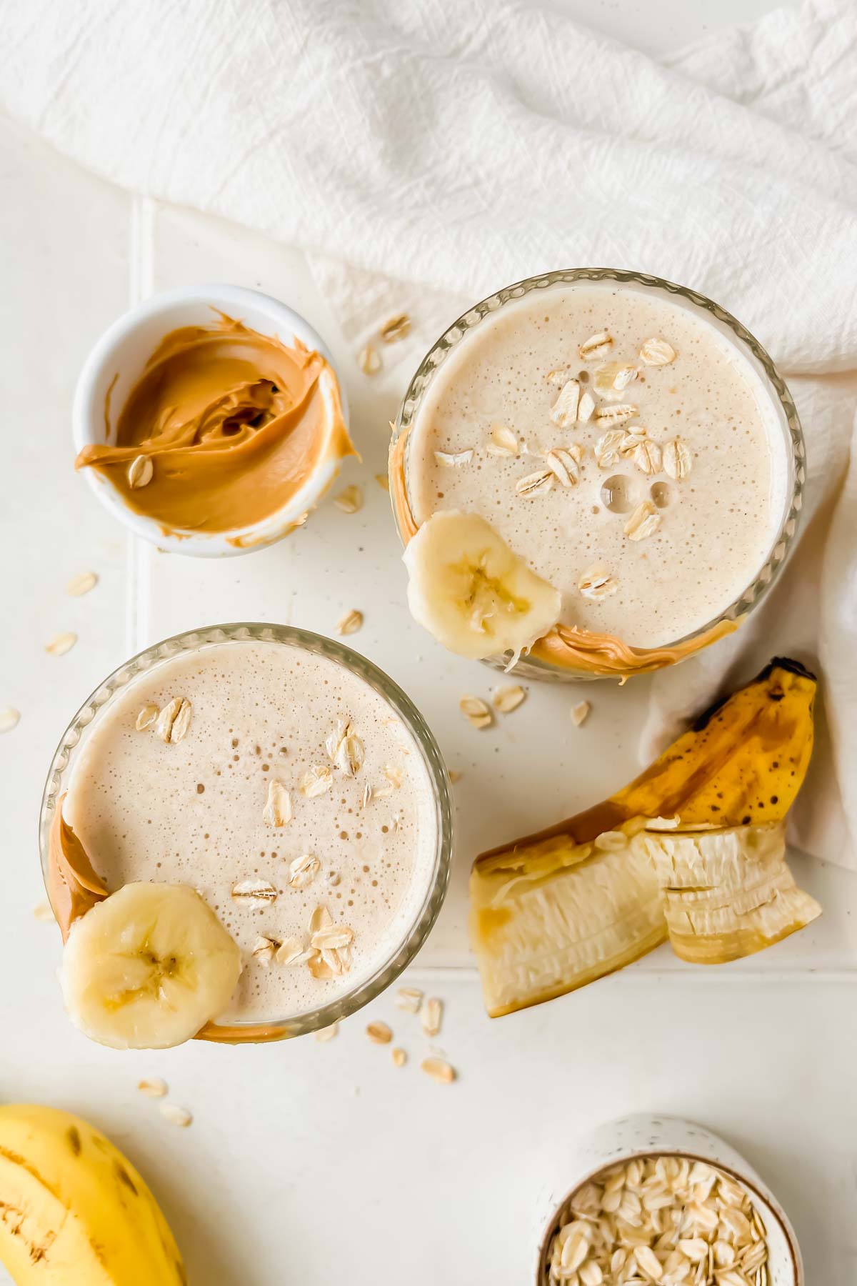 two banana bread protein smoothies in glasses garnished with banana slice, peanut butter and rolled oats.