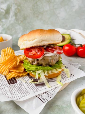 loaded california burger on brioche bun beside ruffle chips on newspaper.