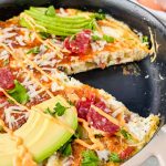 close up of cooked salami and egg frittata topped with avocado and herbs with a slice cut out of it.