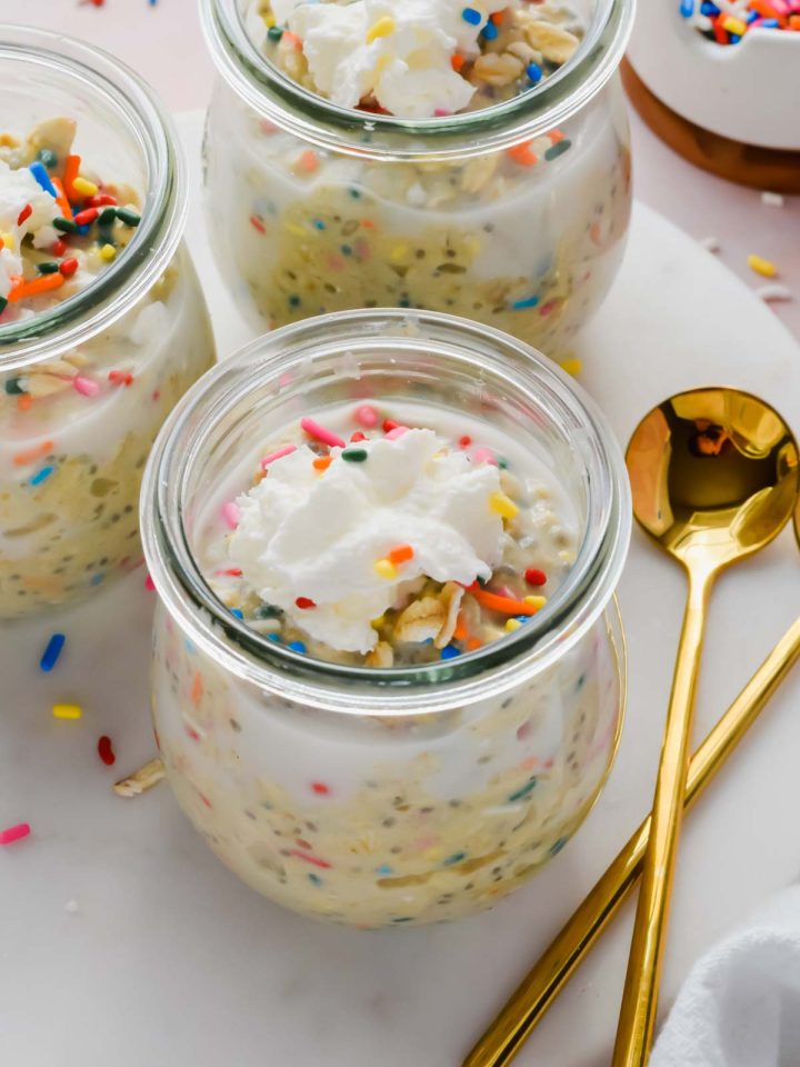 three jars of birthday cake overnight oats topped with whipped cream and rainbow sprinkles beside gold spoons.