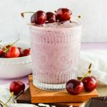 clear glass filled with light pink chocolate cherry smoothie garnished with fresh cherries on a skewer laying across top of glass on wood block with white background and cherries scattered around.