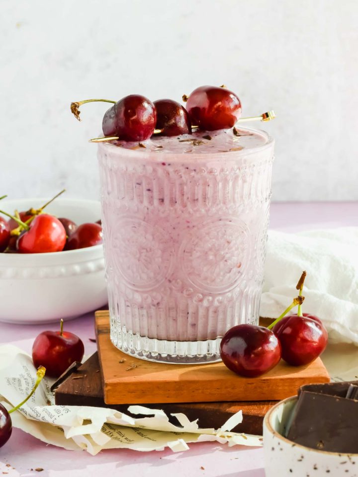 clear glass filled with light pink chocolate cherry smoothie garnished with fresh cherries on a skewer laying across top of glass on wood block with white background and cherries scattered around.