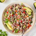 italian quinoa salad topped with avocado slices in ceramic serving bowl with serving spoon in dish.