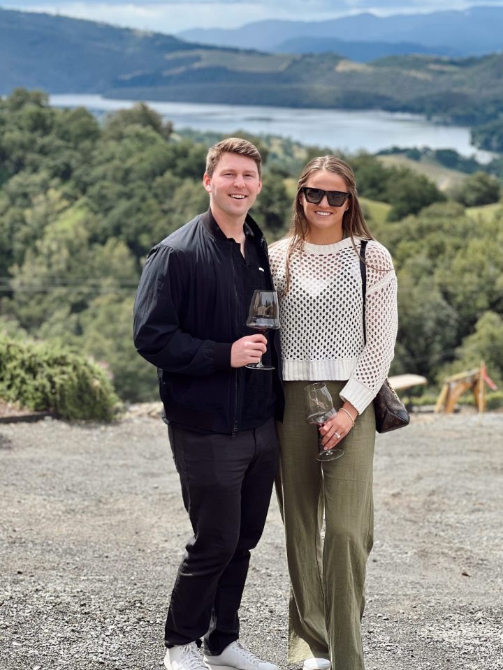 Courtney and jack holding wine glasses with beautiful hills and lake in background.