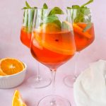3 rosé aperol spritzes in wine glasses garnished with orange slices and mint leaves.
