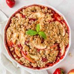 cooked strawberry apple crisp garnished with fresh mint and strawberries on pink tiled backdrop.