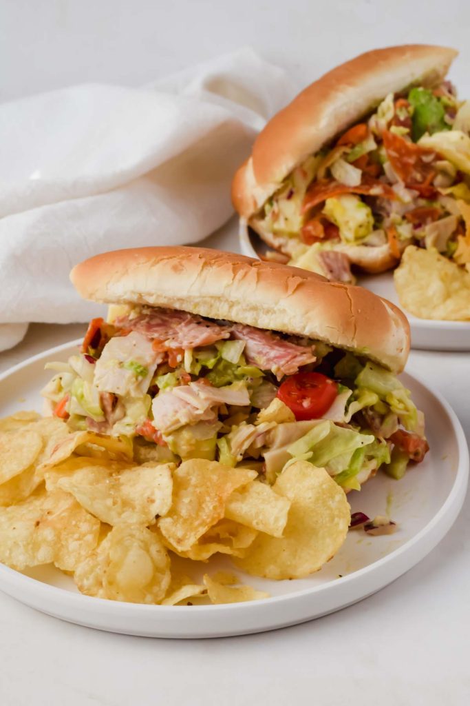 two grinder sandwiches on white plates beside potato chips.