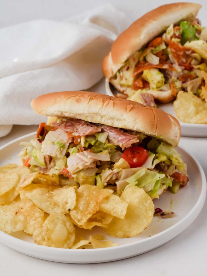two grinder sandwiches on white plates beside potato chips.