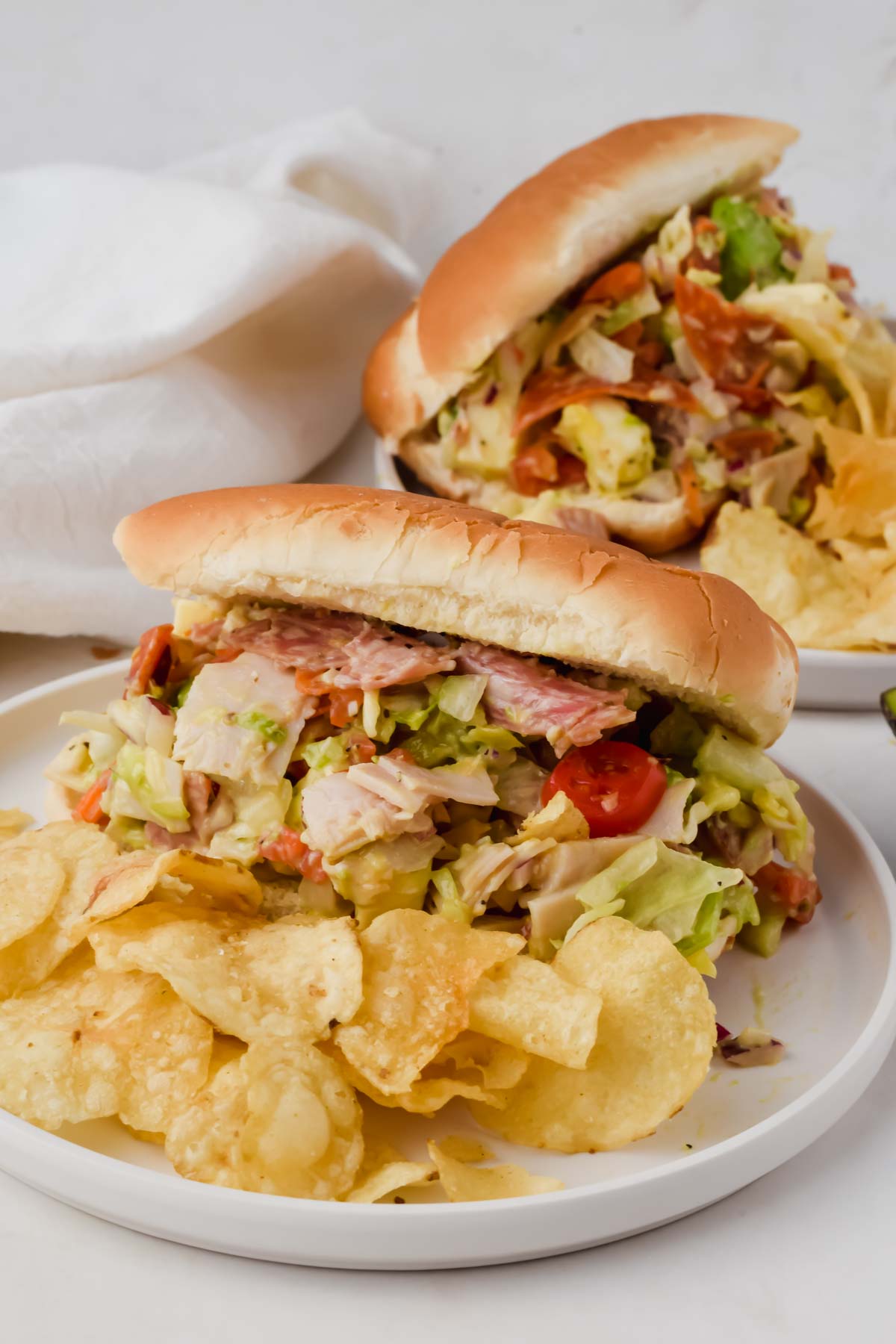 two loaded grinder sandwiches on white plates beside potato chips.