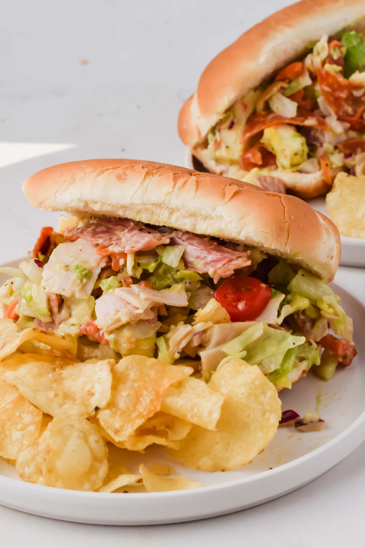two loaded grinder sandwiches on white plates beside potato chips.