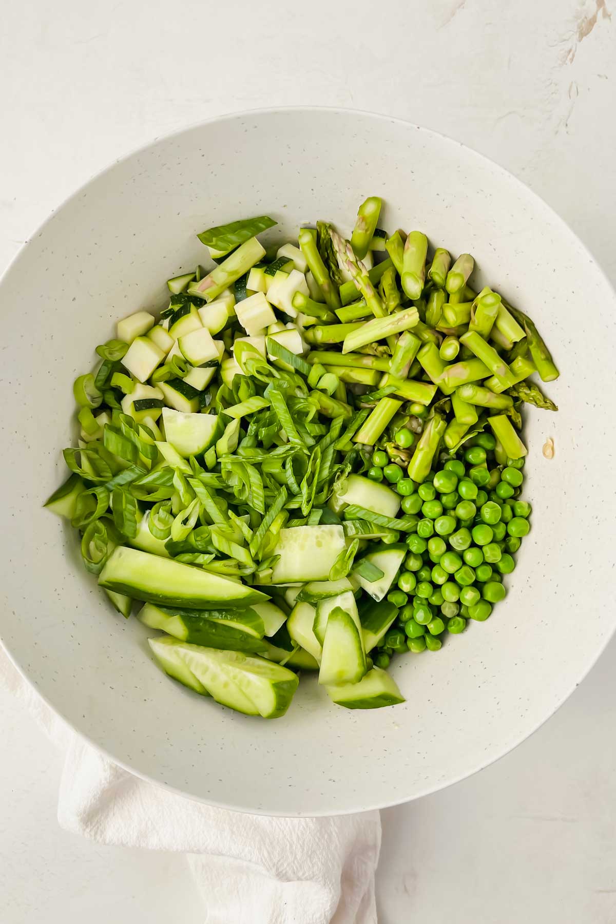 summer greens including cucumbers, peas, zucchini, and asparagus in white mixing bowl.