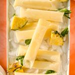 Pina colada ice pops on white tray filled with ice surrounded by pineapple slices.