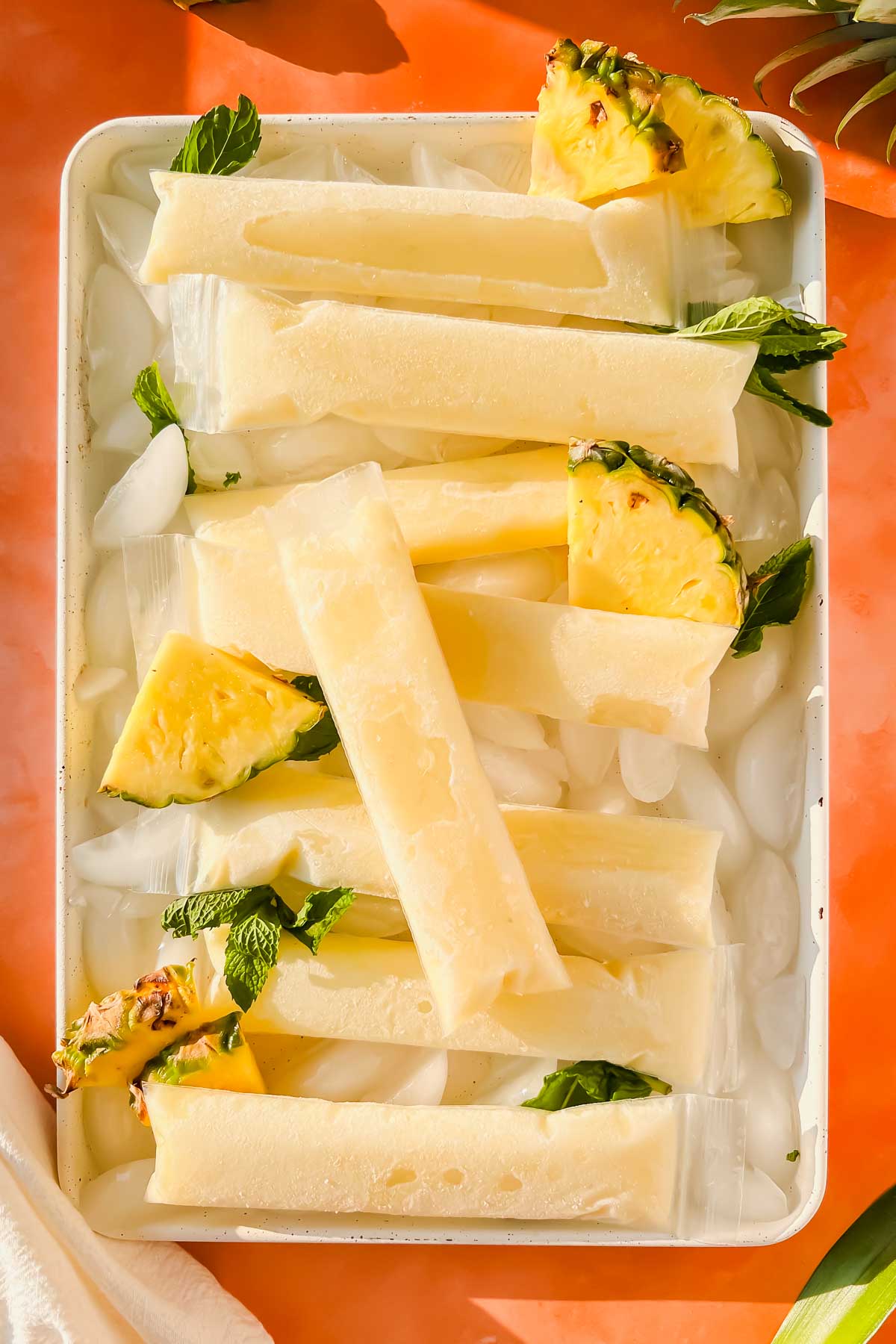 Pina colada ice pops on white tray filled with ice surrounded by pineapple slices.