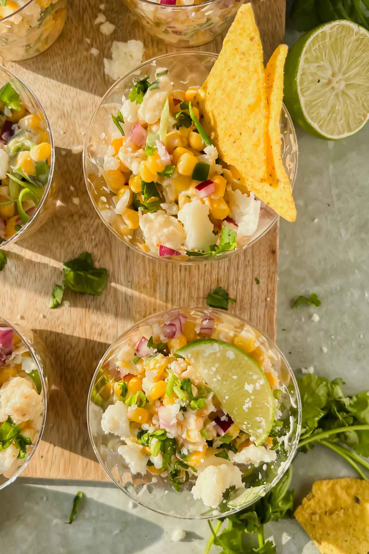 elote in small plastic cups garnished with lime wedges and tortilla chips on wood cutting board.
