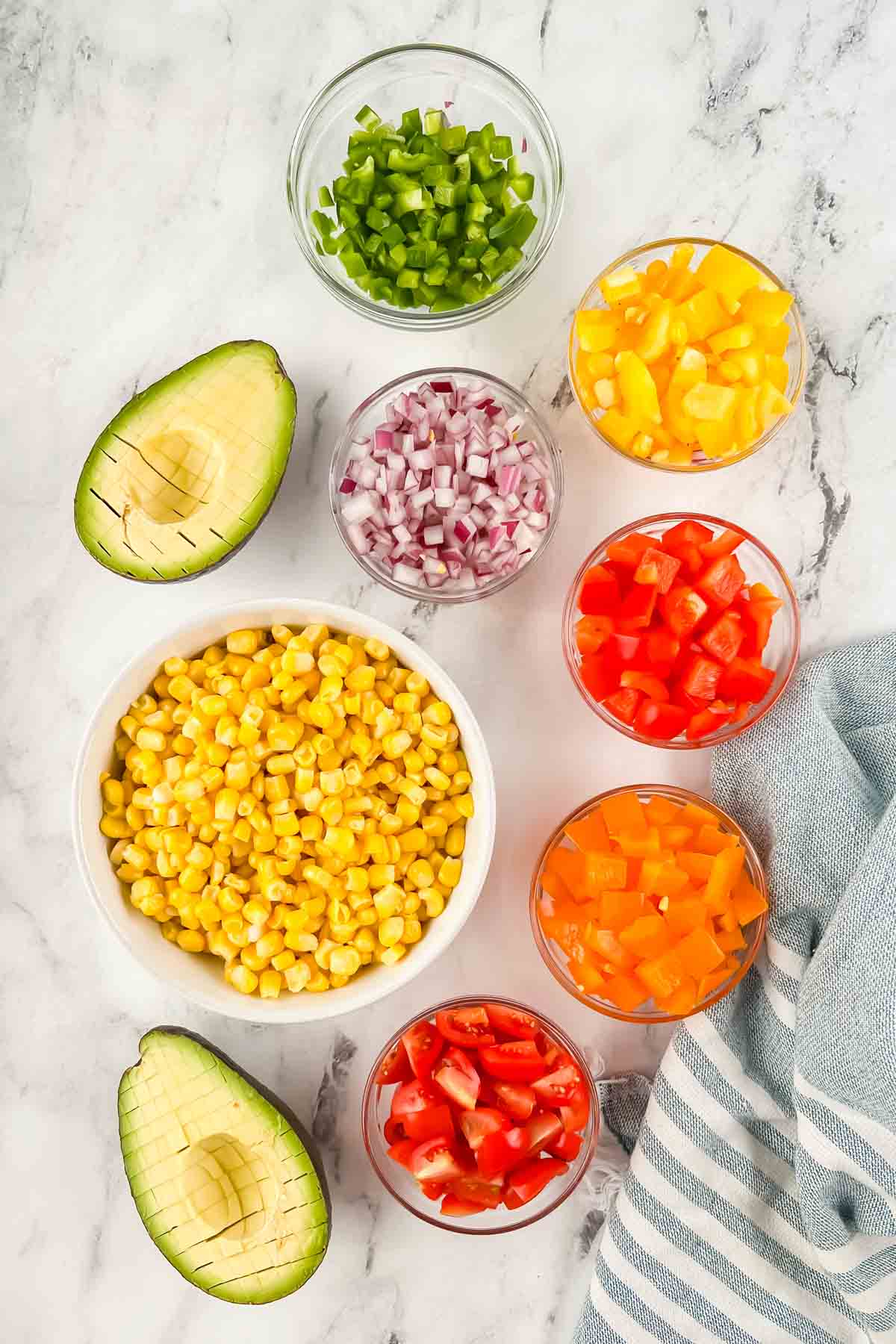 Diced peppers, onion, tomato, corn, and avocado in individual bowls on white marble background