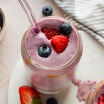 berry smoothie in clear glass with glass straw topped with fresh raspberries and blueberries.