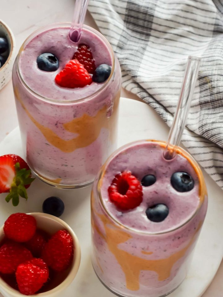 two berry smoothies in clear glasses with glass straws topped with fresh raspberries and blueberries.