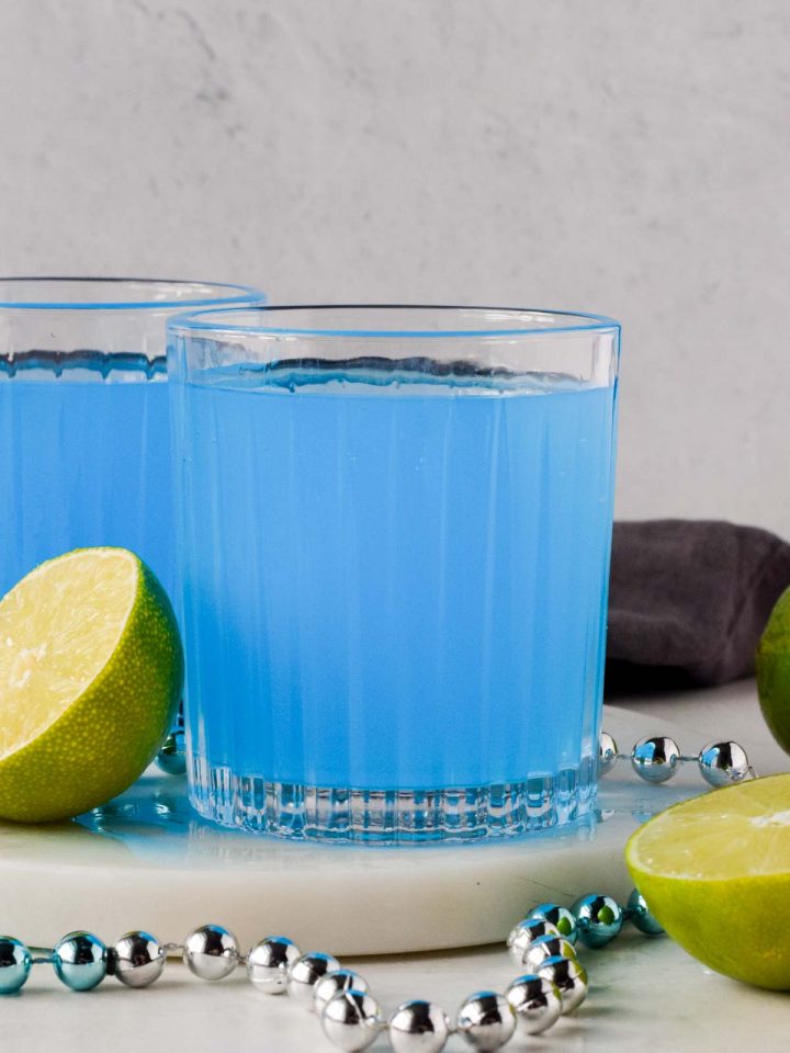 two blue kamikaze drinks garnished with lime and on white marble coaster.