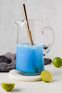 glass pitcher filled with blue kamikaze drink on marble coaster and grey cloth behind it.