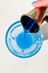 hand pouring a jigger of Blue Curacao into glass cocktail shaker.