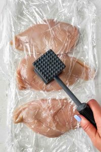 A cutting board is lined with plastic wrap then layered with chicken breasts then more plastic wrap overtop the chicken.