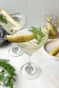two pickle juice martinis garnished with a pickle spear and fresh dill on white background.