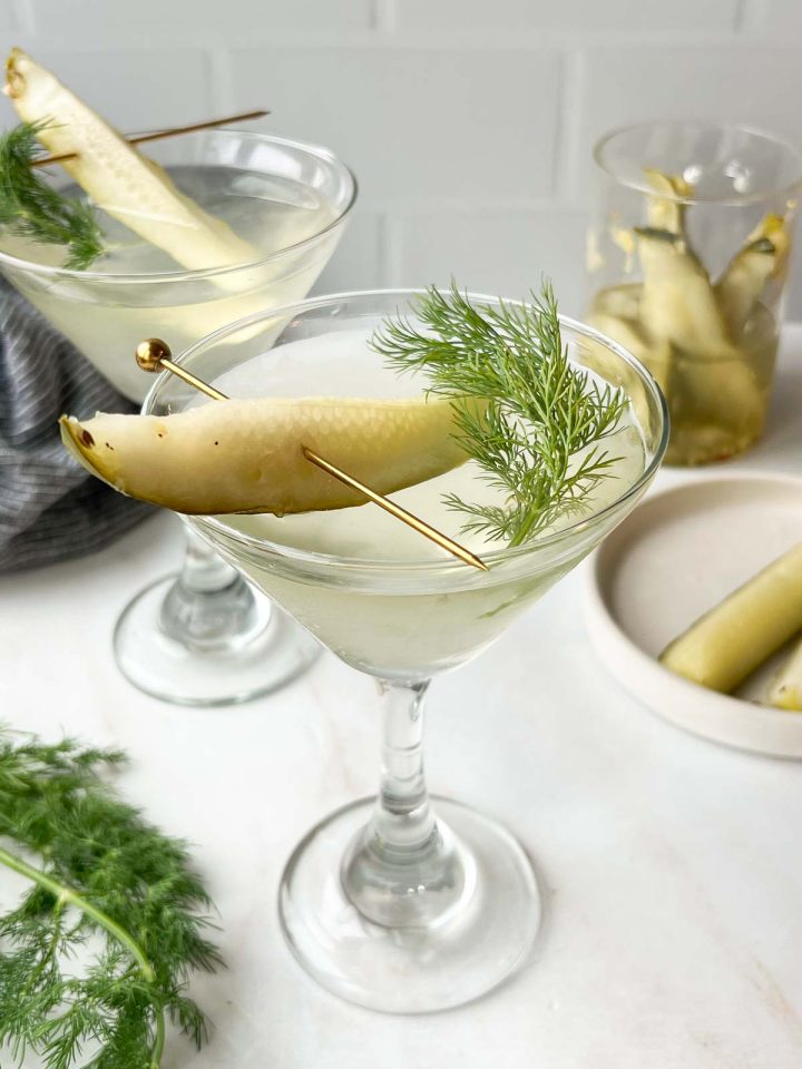 two pickle juice martinis garnished with a pickle spear and fresh dill on white background.