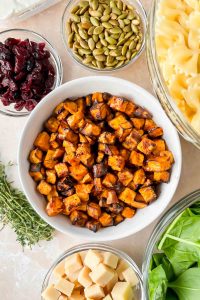 roasted sweet potato cubes in white bowl surrounded by additional ingredients for fall pasta salad.