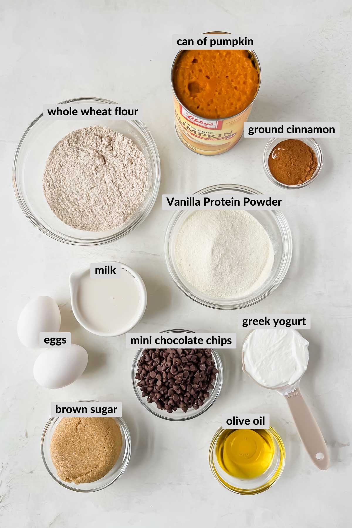 labeled ingredients for pumpkin protein muffins in individual bowls on white background.