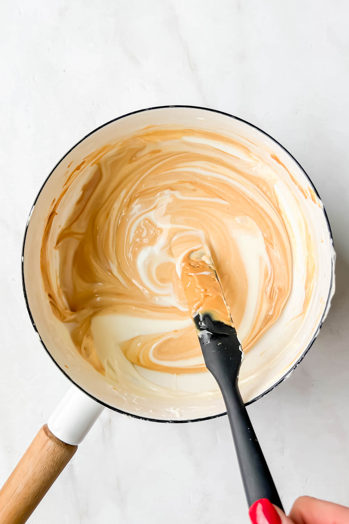 Overhead view of melted white chocolate and peanut butter in sauce pot with rubber scraper.
