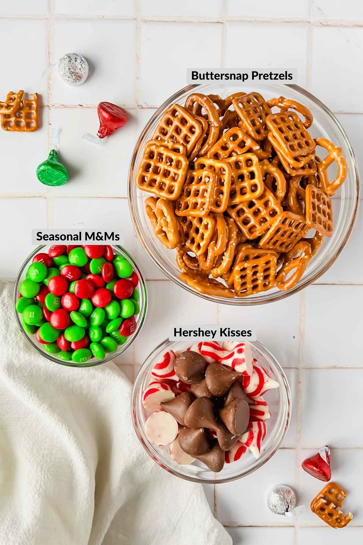 ingredients for pretzel hershey M&Ms in individual bowls on white tile background.