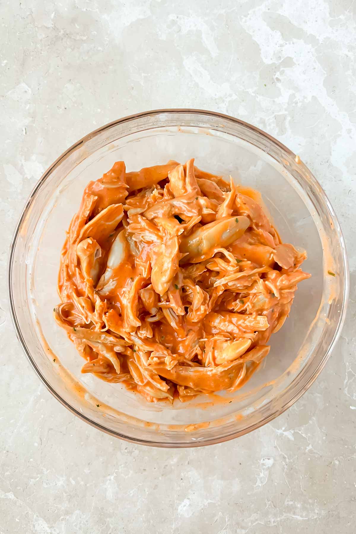 shredded chicken covered in buffalo sauce in glass mixing bowl.