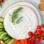 cottage cheese ranch dip with fresh dill as garnish on plate with cucumbers, carrots and pretzels.