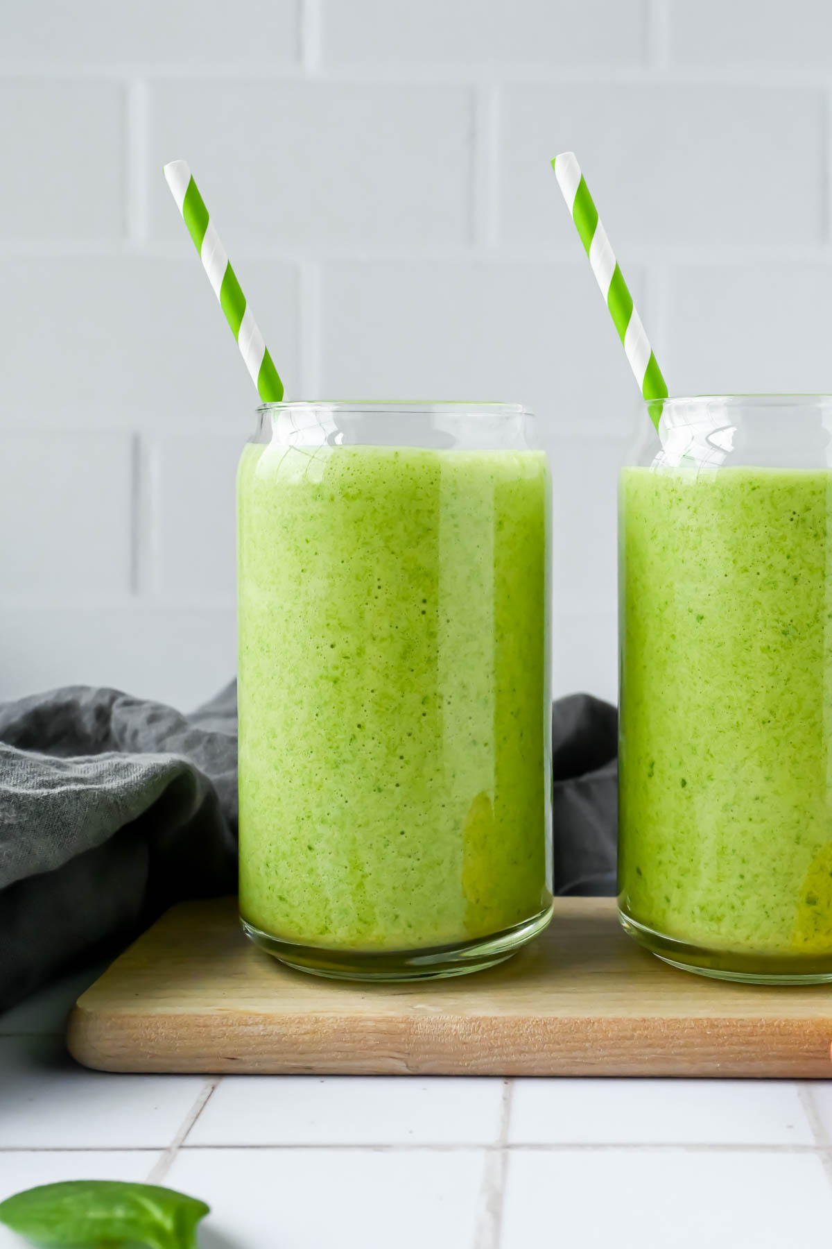 two mango spinach smoothies with a green and white striped straw on white tile background. 