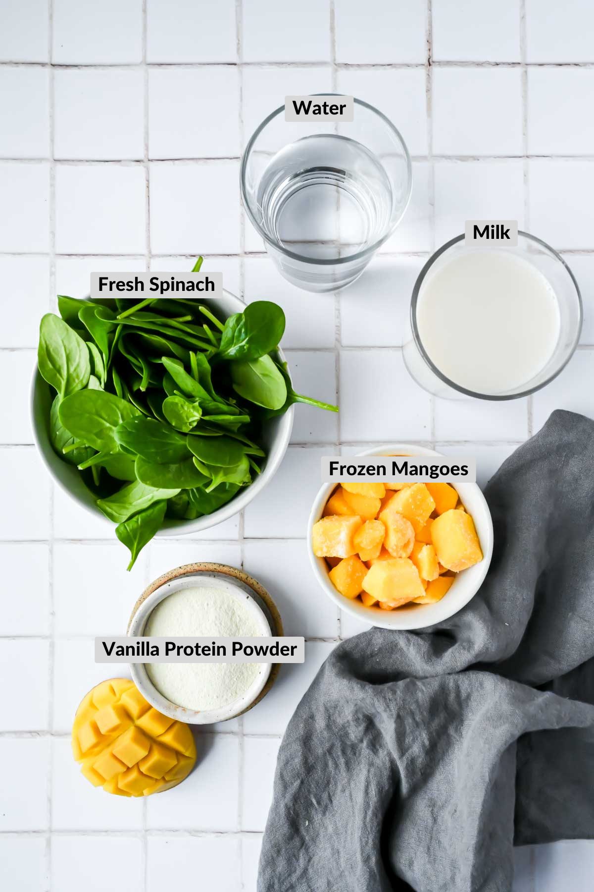 ingredients for mango spinach protein smoothie in individual bowls on white tiled background.