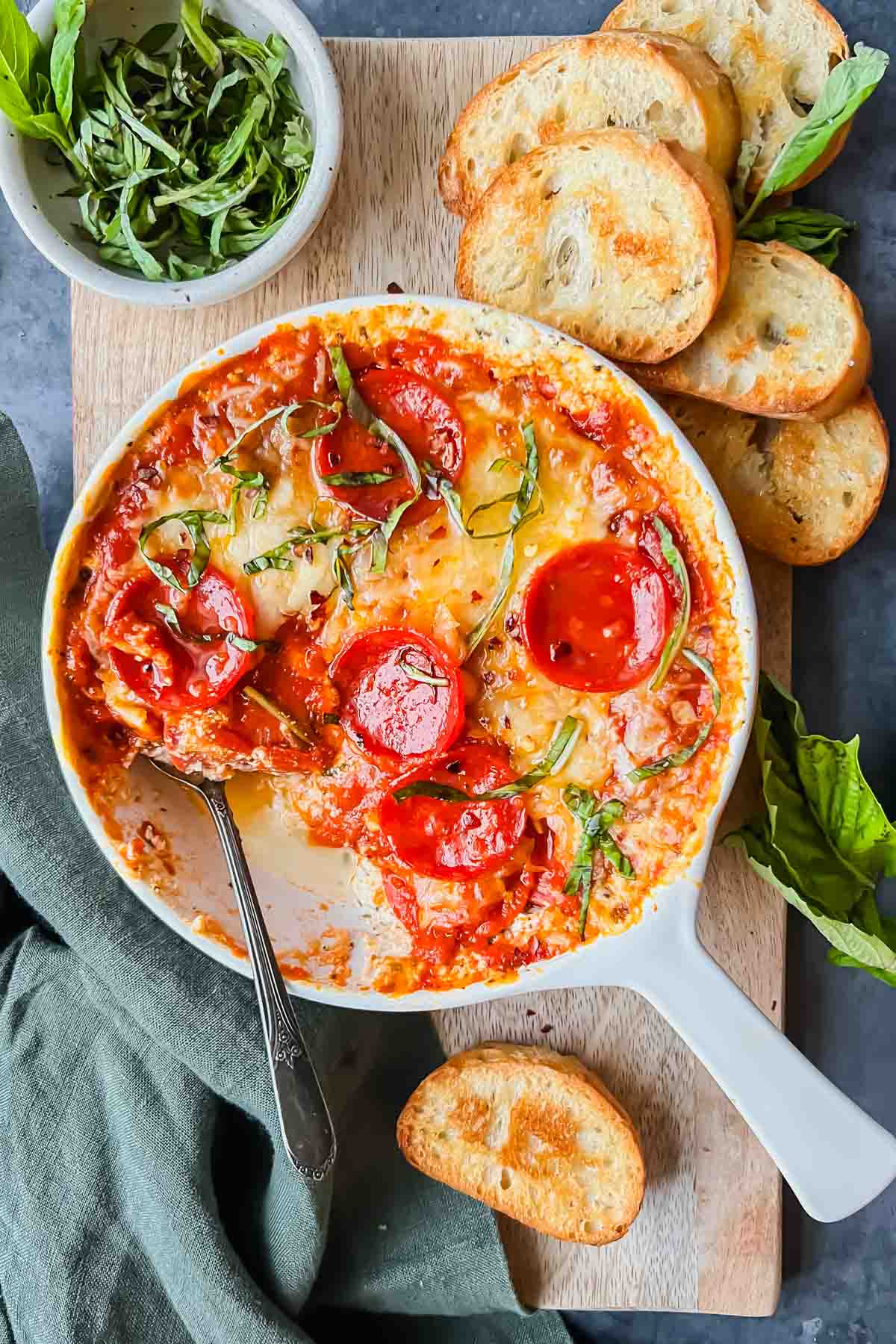 baked pepperoni pizza dip in white skillet garnished with fresh basil on serving board with baked crostinis.