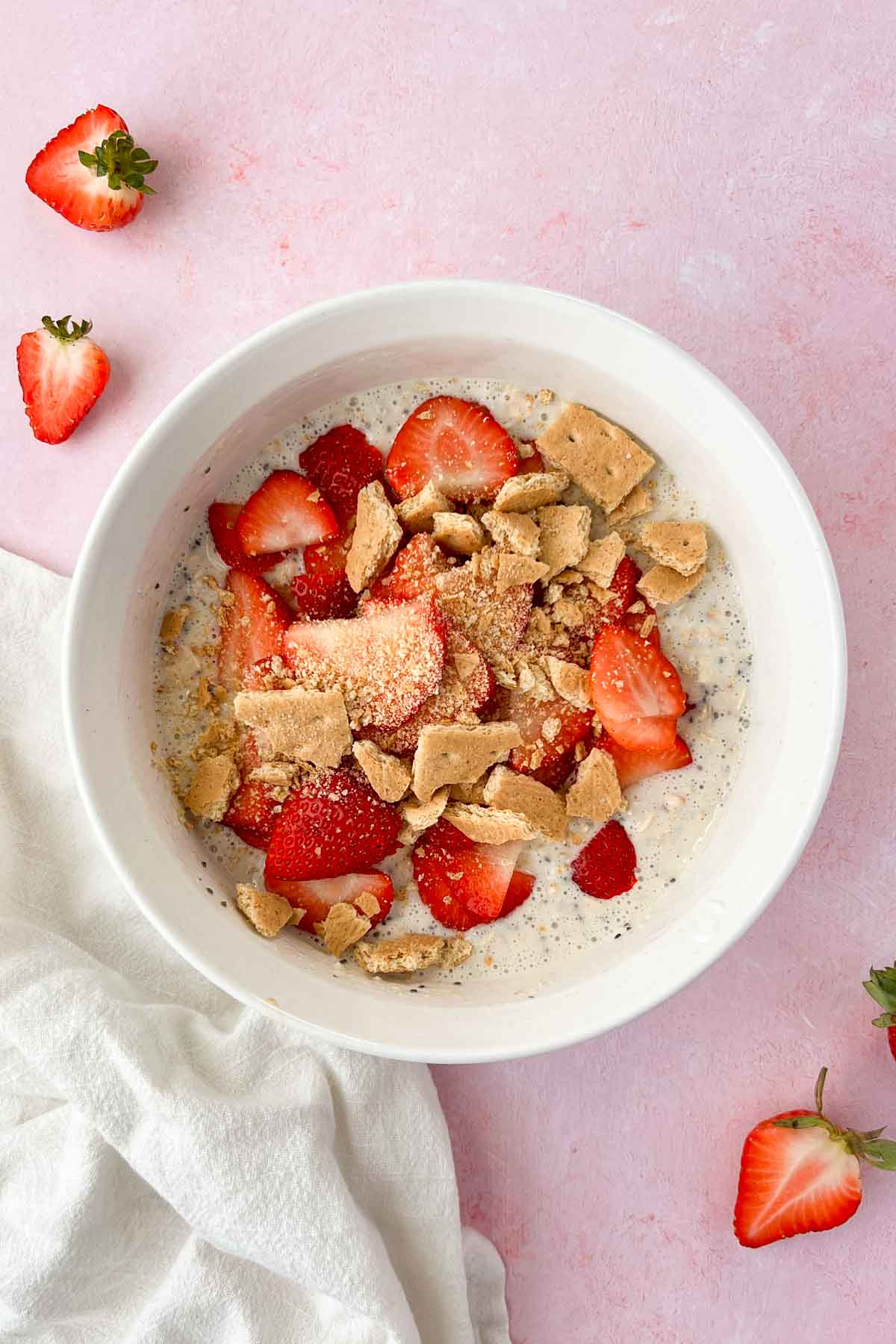 strawberry cheesecake oats ingredients mixed in white bowl with extra strawberries and graham crackers on top.