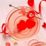 two heart shaped alcohol gummies in a coupe glass topped with rosé and garnished with red ribbons.