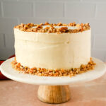 frosted carrot cake garnished with crushed walnuts on marble cake stand.