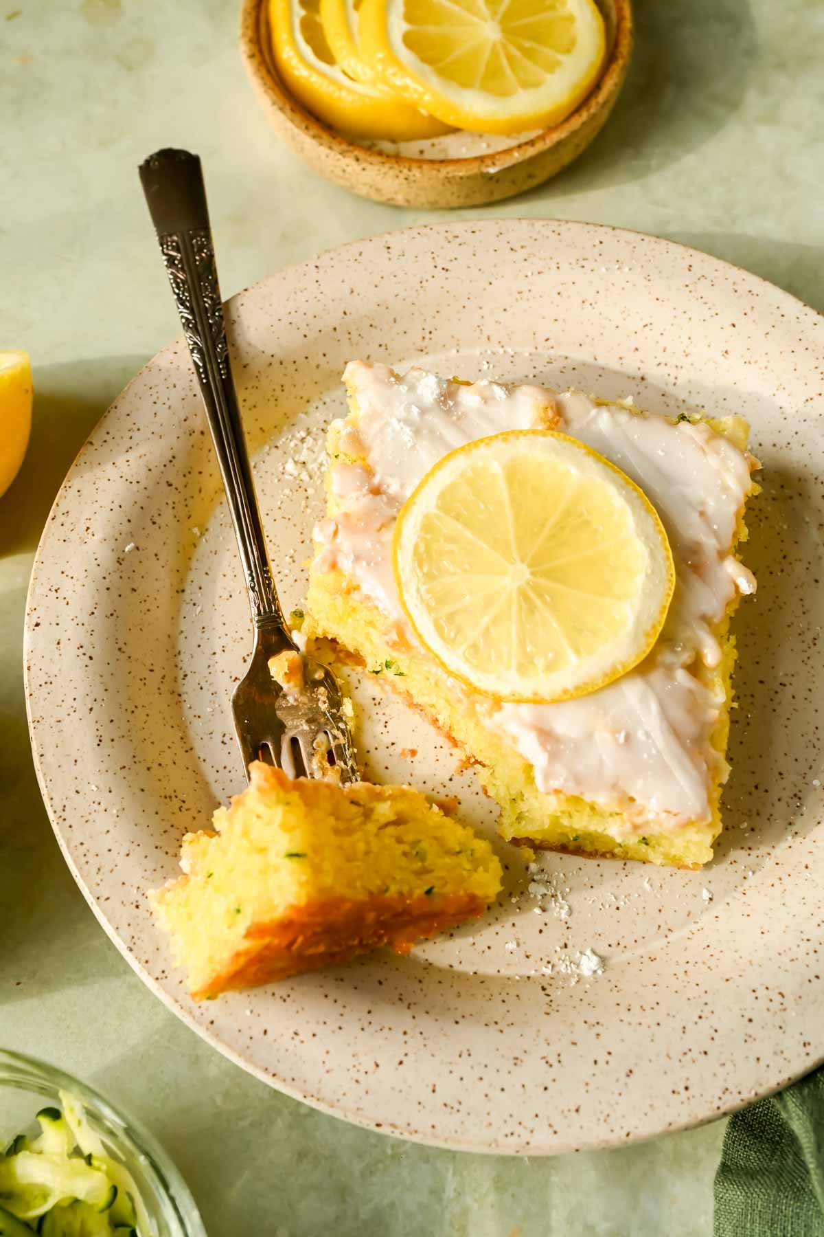 slice of lemon zucchini cake glazed with lemon slice on top on speckled plate.