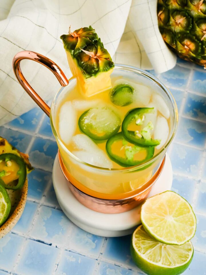 spicy pineapple moscow mule garnished with fresh pineapple and jalapeno slices on blue tiled backdrop.